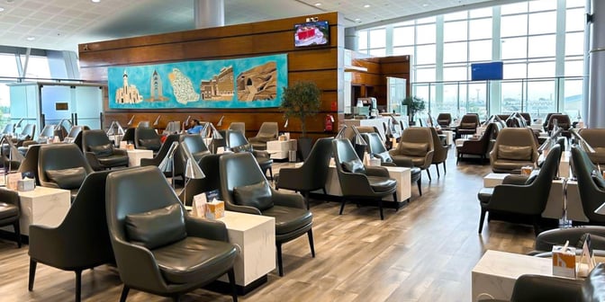 The seating area at Hayyak Lounge features comfortable chairs with individual tables, set against large windows offering airport views.