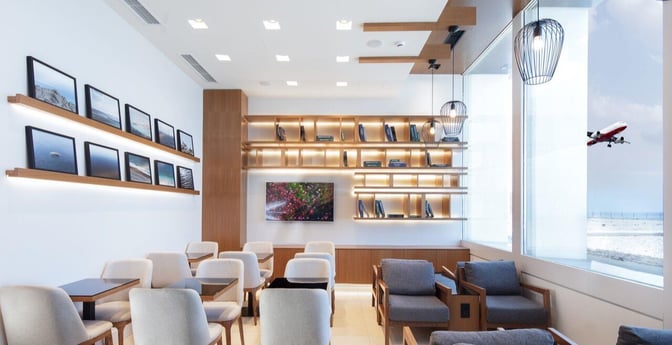 Seating area in Goldair Handling Lounge at Heraklion Airport features tables, chairs, and wall shelves with books and framed photos.