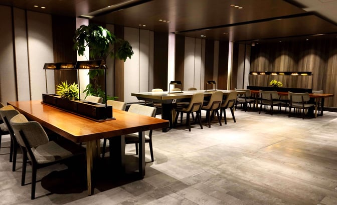 The seating area in Plaza Premium First lounge at Hong Kong International Airport features communal tables with chairs and decorative plants.