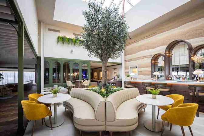 The No1 Lounge at Jersey Airport features a seating area with round tables, cushioned chairs, and a central decorative tree.