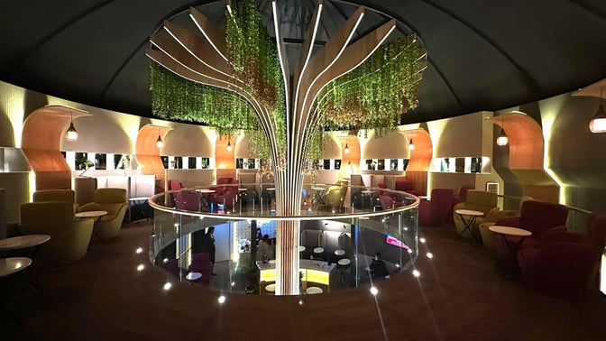 The Sky Majestic Lounge at Jinnah International Airport features a circular seating area with armchairs and tables, surrounded by decorative greenery.