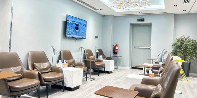The seating area in Hayyak Lounge at King Abdullah bin Abdulaziz International Airport features comfortable chairs, small tables, and a wall-mounted TV.