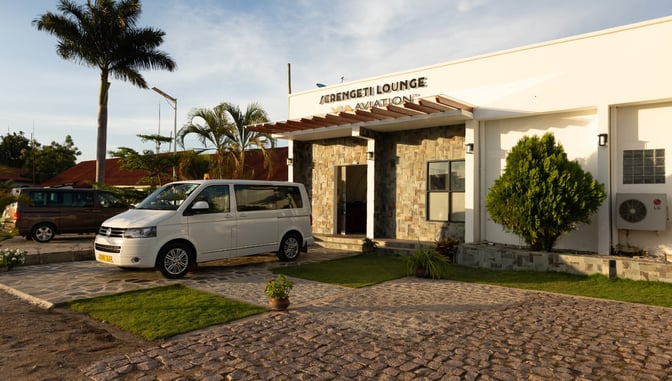 The entrance to Serengeti Lounge at Julius Nyerere International Airport features a stone facade and a driveway with parked vehicles.