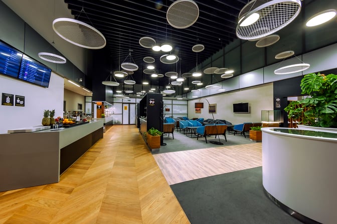 The Krakow Airport Business Lounge features a seating area with blue chairs, a food counter, and flight information screens.