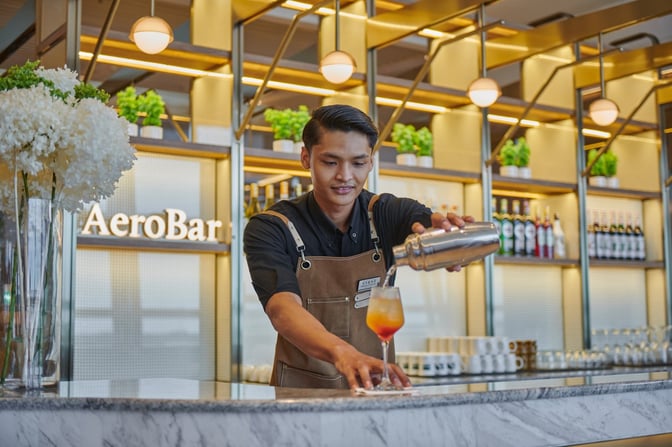 The AeroBar at Plaza Premium Lounge, Kuala Lumpur International Airport, features a bartender preparing a cocktail with various spirits displayed.