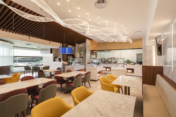 The Travel Club Lounge at Kuala Lumpur International Airport features a dining area with marble tables, diverse seating, and self-serve food stations.