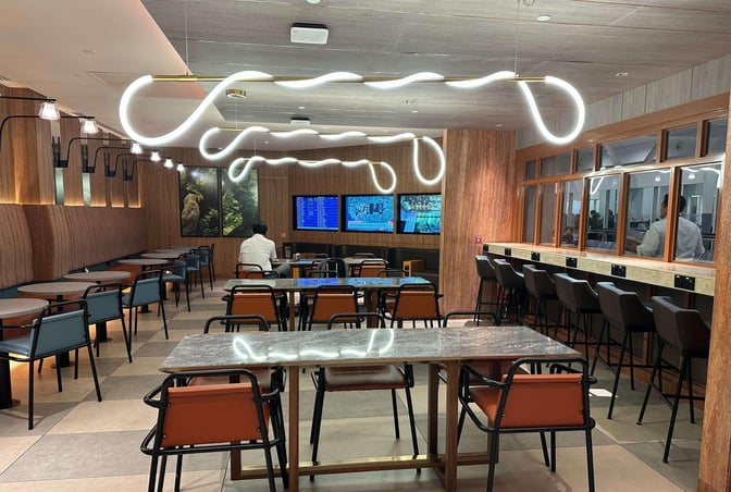 The seating area in the Travel Club Lounge at Kuala Lumpur International Airport features tables, chairs, and flight information screens.