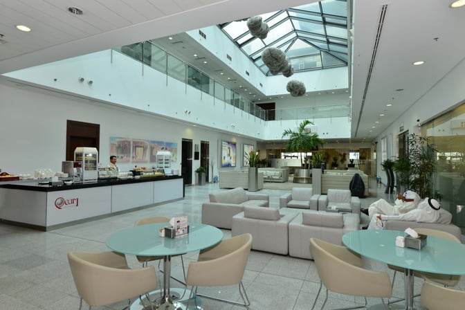 The Pearl Lounge at Kuwait International Airport features a seating area with tables and chairs, adjacent to a self-serve food counter.