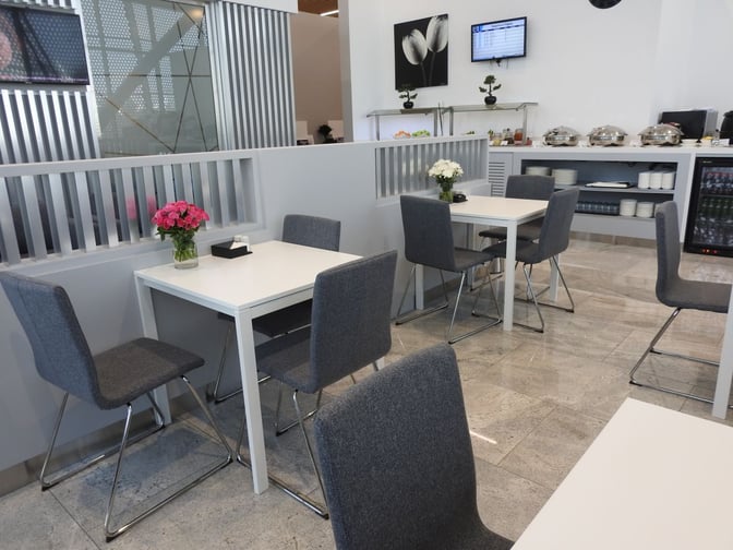 Seating area in Pearl Lounge at Kuwait International Airport features tables with chairs and a self-serve food station in the background.
