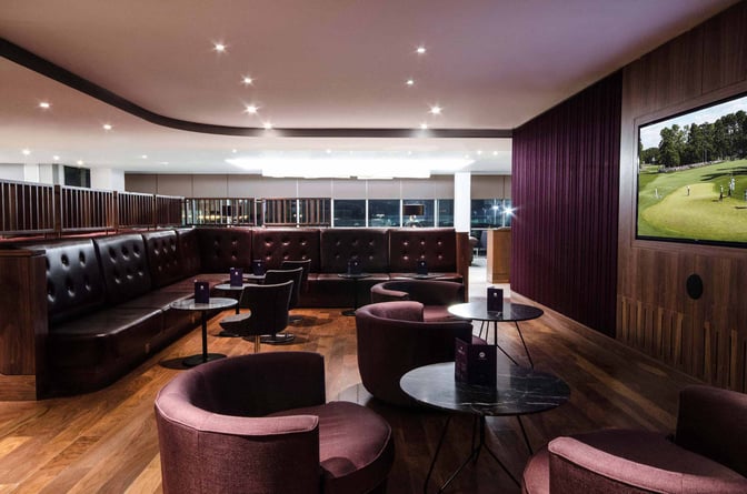 The No1 Lounge at London Gatwick Airport features a seating area with round tables, cushioned chairs, and a wall-mounted TV.
