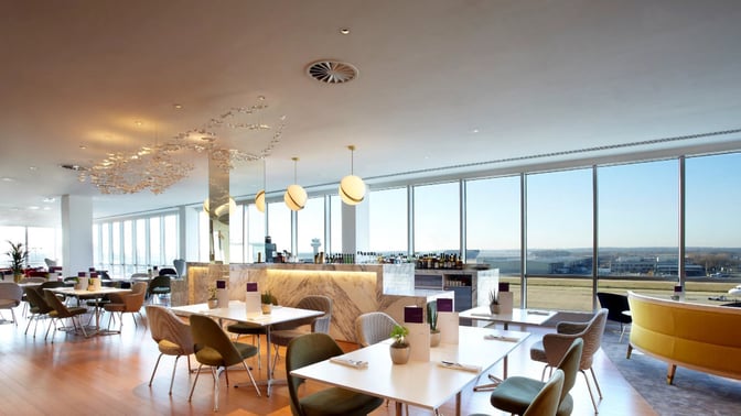 The Plaza Premium Lounge at London Gatwick Airport features a seating area with tables and chairs, offering views of the runway.