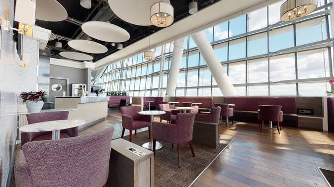 The Club Aspire Lounge at London Heathrow Airport features a seating area with tables, chairs, and large windows offering airport views.