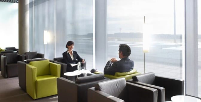 Seating area at Luxembourg Findel Airport's lounge features armchairs and small tables, offering views of the airport through large windows.
