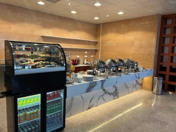 The food area at Pearl Lounge, Luxor International Airport, features a buffet with chafing dishes and a refrigerated display for beverages.
