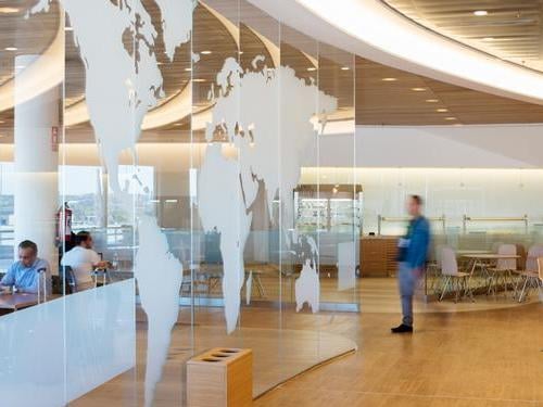 The seating area in Sala Cibeles at Madrid Barajas Airport features tables and chairs with a world map glass partition.