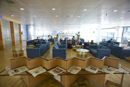 The seating area in Sala Puerta Del Sol at Madrid Barajas Airport features comfortable chairs and tables with available newspapers for guests.
