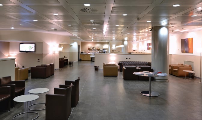 The Monteverdi Classic Lounge at Malpensa Airport features a seating area with armchairs and tables, and a bar offering beverages.
