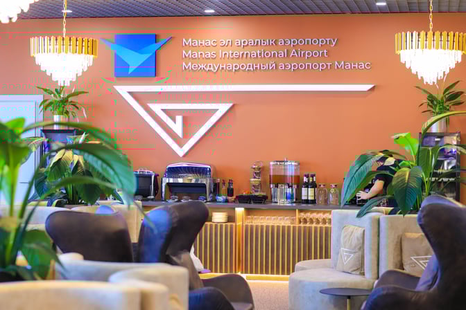 The Manas International Airport business lounge features a self-serve beverage station with seating and decorative plants for relaxation.