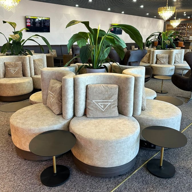 Circular seating arrangements in the Manas International Airport Business Lounge offer comfortable spaces with small side tables and decorative plants.