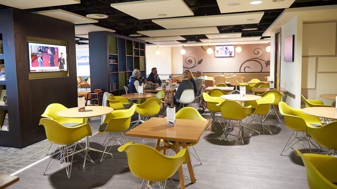 The seating area in Manchester Airport's Escape Lounge features round tables and chairs, with a television for entertainment.