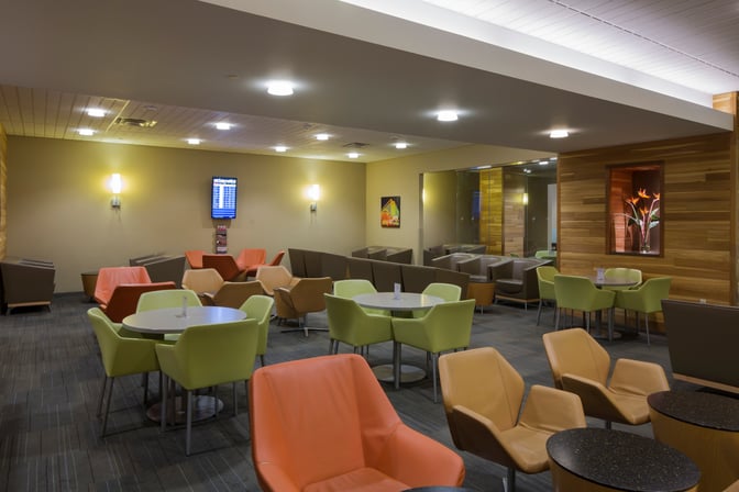 The seating area at The Club LAS features various chairs and tables, with a flight information screen for travelers.