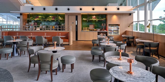 The Plaza Premium Lounge at Kualanamu International Airport features a seating area with tables and chairs near a self-serve food station.