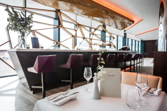 The House lounge at Melbourne Airport features a bar with high seating and a dining table set with glassware and cutlery.