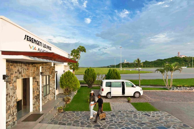The entrance to Serengeti Lounge at Mwanza Airport features a stone facade and is adjacent to a parking area with a van.