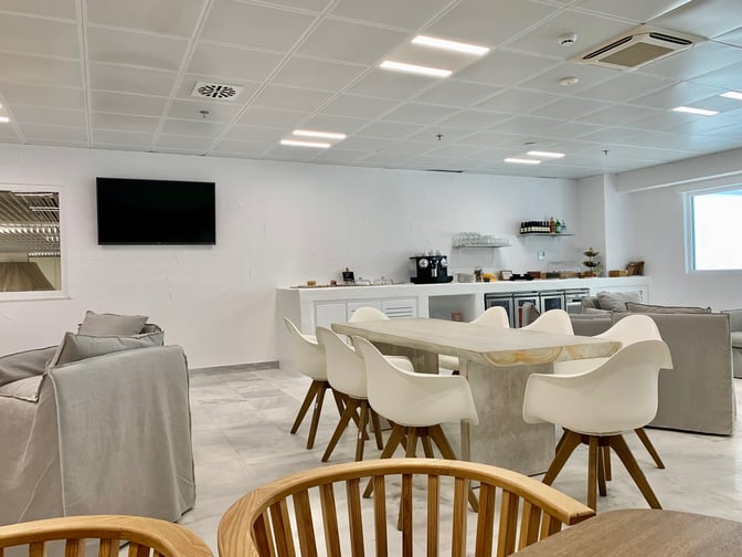 The lounge at Mykonos Island National Airport features a seating area with a communal table, sofas, and a self-serve refreshment counter.