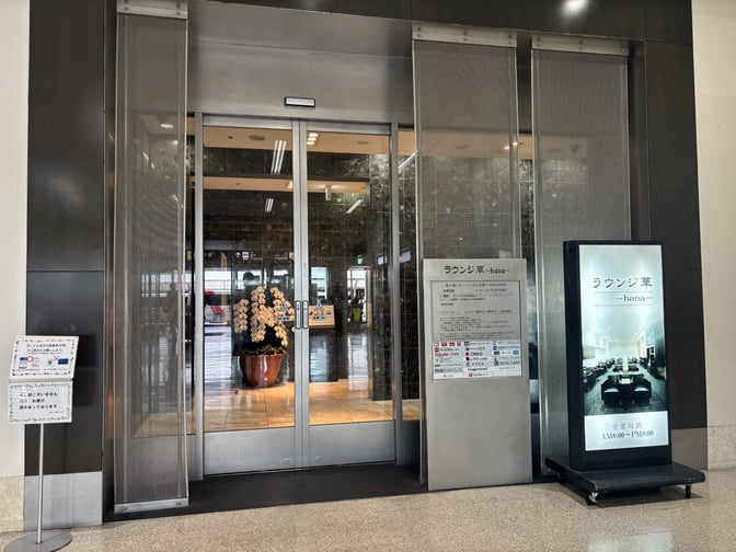 The entrance to Lounge Hana at Naha Airport features sliding glass doors and informational signage displaying operating hours and amenities.