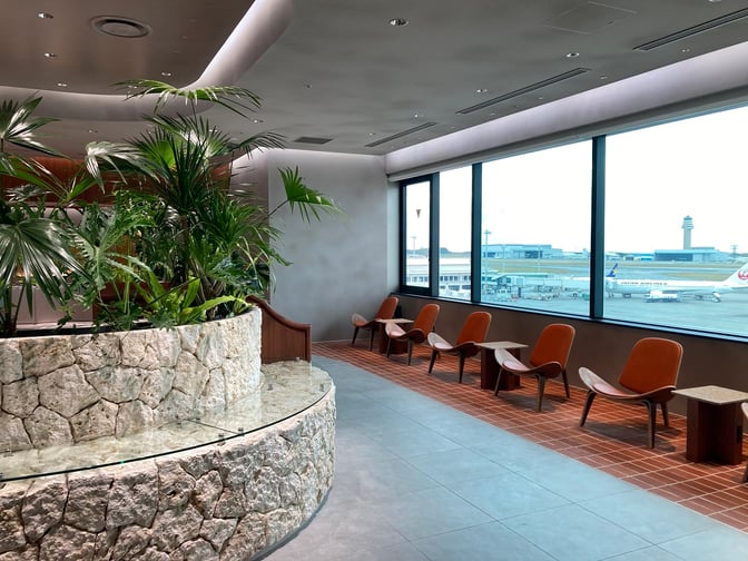 The seating area at Lounge Hana, Naha Airport, features chairs with tables overlooking the runway through large windows.