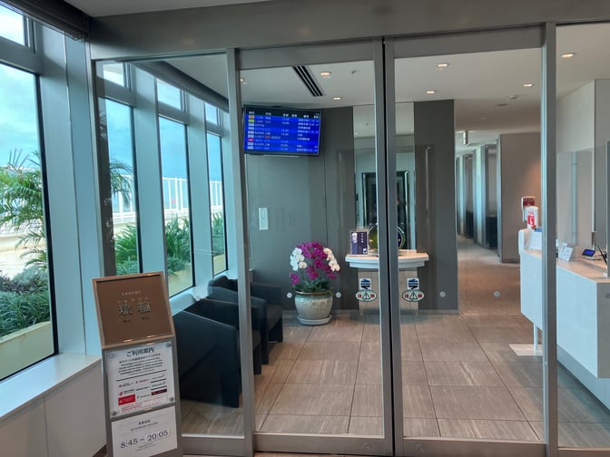 The entrance to Lounge RYURIN at Naha Airport features glass doors, seating, and flight information displays near the reception area.