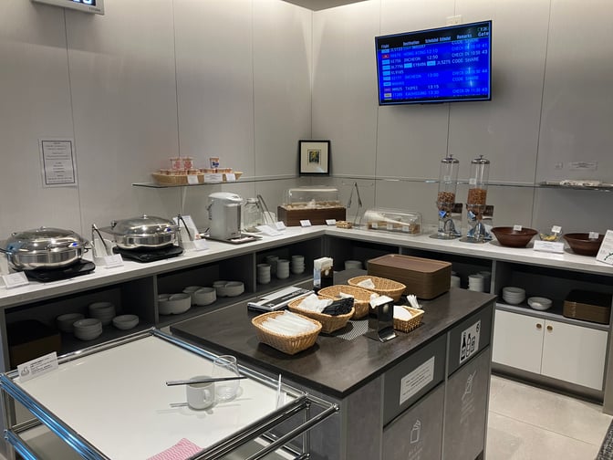 The self-serve food area at Lounge RYURIN, Naha Airport, offers hot dishes, snacks, and beverages with flight information displayed above.