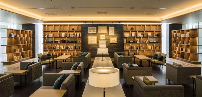 The seating area at CAFÉ SKY LIBRARY in New Chitose Airport features comfortable chairs and tables surrounded by bookshelves.