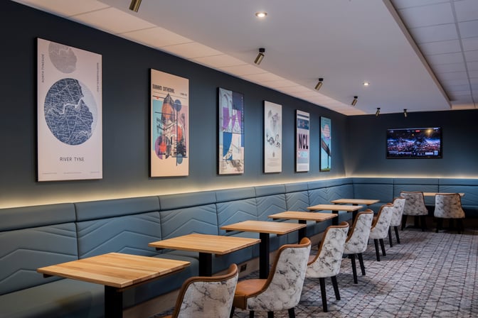 The Aspire Lounge at Newcastle Airport features a seating area with tables, chairs, and wall art, providing a comfortable space for travelers.