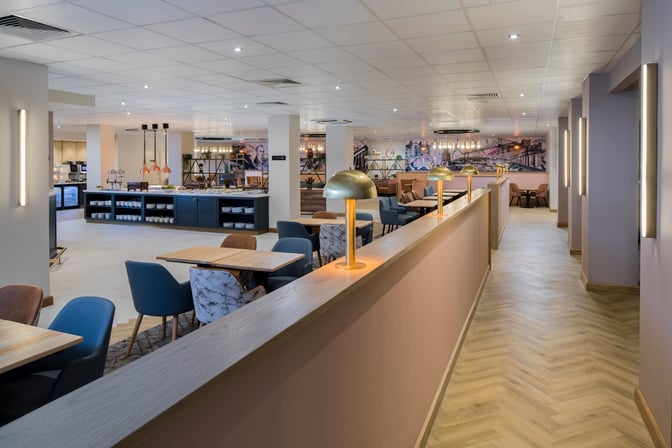 The Aspire Lounge at Newcastle Airport features a spacious seating area with tables and chairs, adjacent to a self-serve food station.
