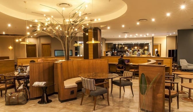 The Aspire Lounge at O. R. Tambo International Airport features a seating area with tables and chairs, adjacent to a staffed bar.