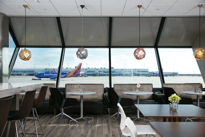 The seating area in the Escape Lounge at Oakland International Airport offers tables with airport runway views through large windows.