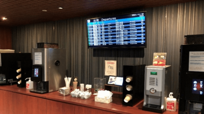 The beverage station at Lounge Osaka in Osaka-Itami Airport features coffee machines, cups, and a departures screen for travelers.