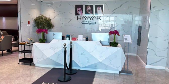 The reception area of Hayyak Lounge at Prince Abdul Mohsin bin Abdulaziz International Airport features a check-in desk and seating area.