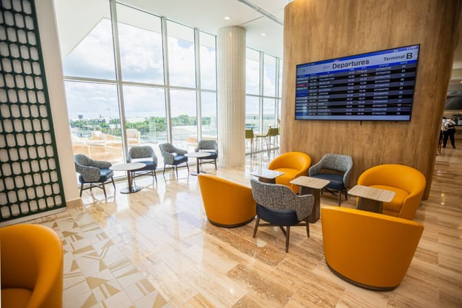 Seating area in Punta Cana International Airport's VIP Lounge features comfortable chairs, tables, and a departures screen with airport views.