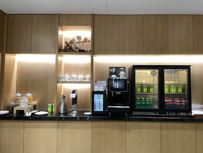 The self-serve beverage station at Plaza Premium Lounge Qingdao offers coffee, tea, and chilled drinks with cups and snacks available.