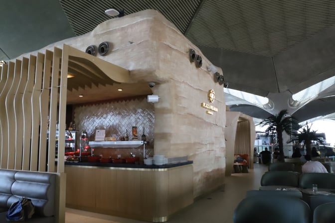 The Plaza Premium Lounge at Queen Alia International Airport features a food service area with self-serve stations and seating nearby.