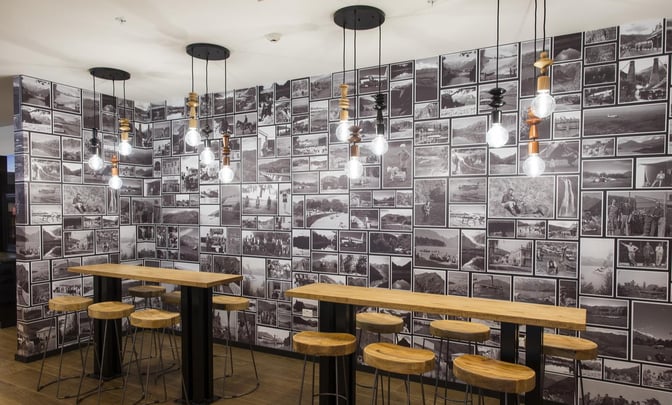 High-top tables with stools are set against a photo collage wall in the Manaia Lounge at Queenstown Airport.