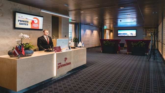 The reception area of the Primeclass Business Lounge at Riga International Airport features a staffed desk and seating area with a screen.