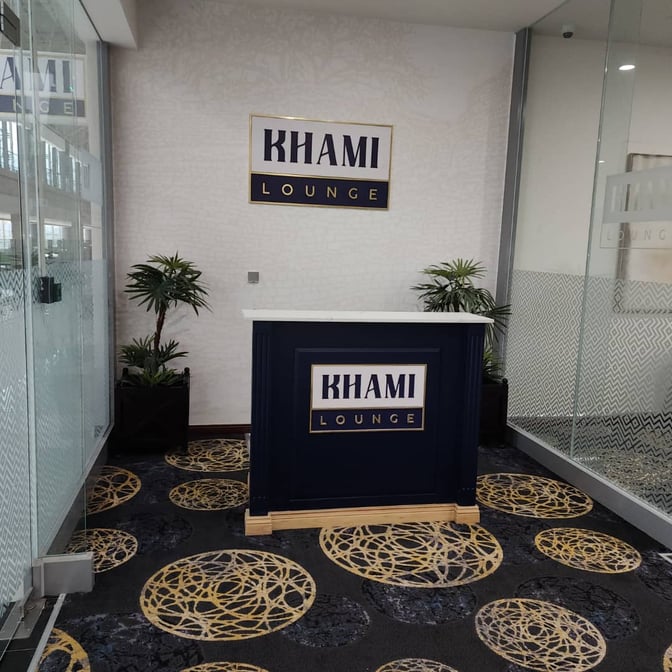 The entrance to Khami Lounge at Robert Mugabe International Airport features a reception desk and signage, with decorative plants nearby.
