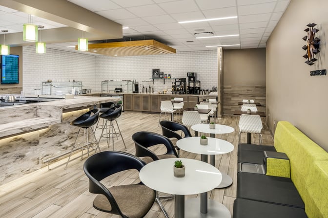 The Escape Lounge at Sacramento International Airport features a seating area with tables and chairs, adjacent to a self-serve food station.