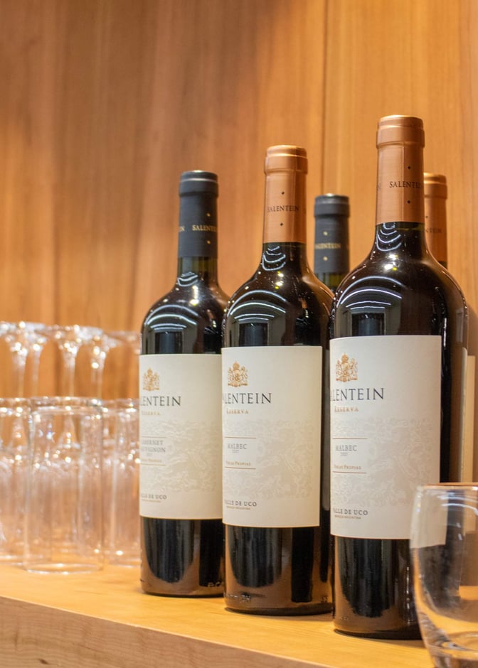 Wine bottles and glasses are displayed on a wooden shelf at the Bariloche AMAE Lounge, San Carlos de Bariloche Airport.