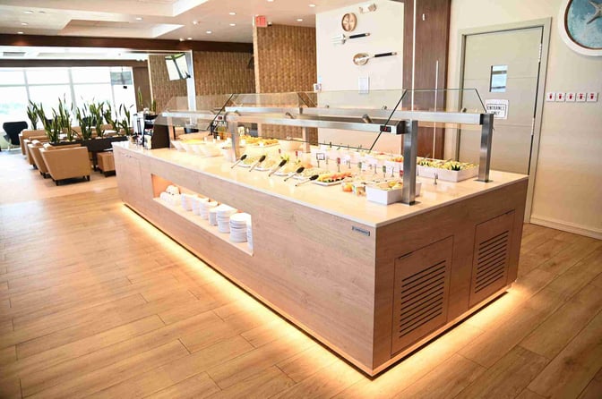 The Club Mobay Departures Lounge at Sangster International Airport features a self-serve buffet with various food options and seating nearby.