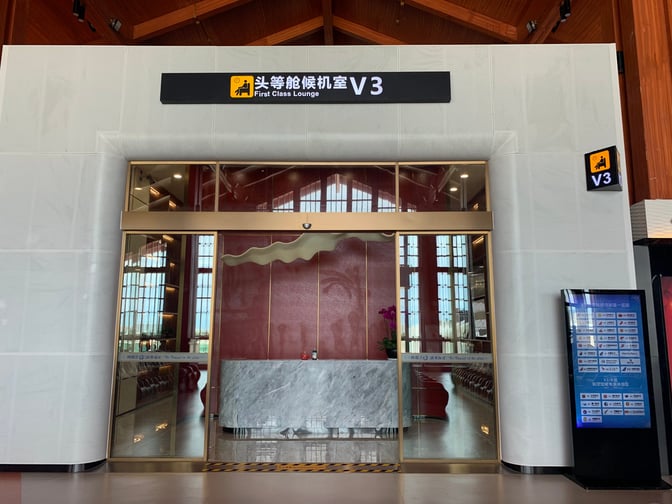 The entrance to the V3 First Class Lounge at Sanya Phoenix International Airport features glass doors and a digital information screen.
